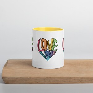 May include: White ceramic mug with a yellow rim. The mug features a colorful graphic of a heart with the words "Love is Love" inside.
