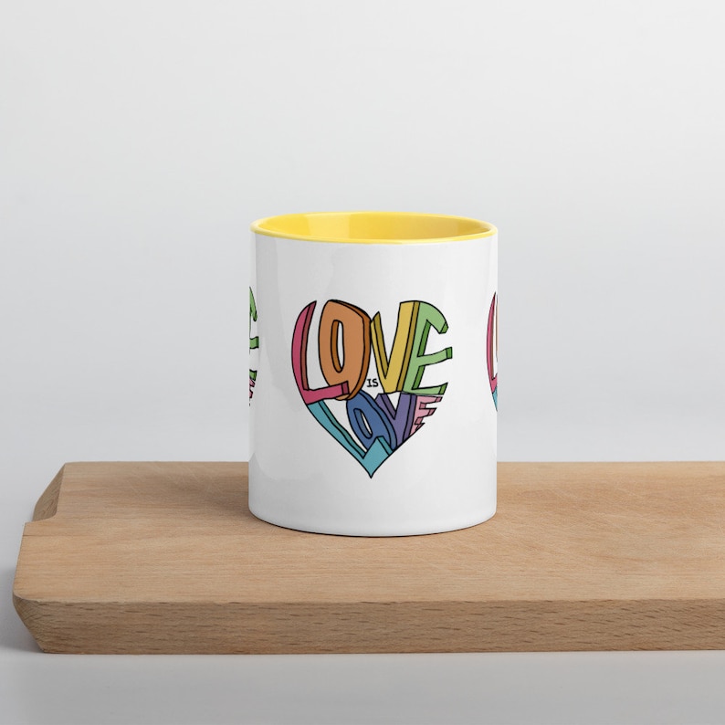 May include: White ceramic mug with a yellow rim. The mug features a colorful graphic of a heart with the words "Love is Love" inside.