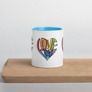 May include: White ceramic mug with a light blue rim. The mug features a colorful graphic of a heart with the words "Love is Love" inside.
