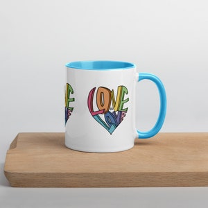 May include: White ceramic mug with a blue rim and handle. The mug features a colorful graphic of the word "LOVE" in a heart shape. The text "LOVE IS LOVE" is written inside the heart.