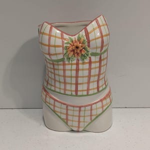 May include: A ceramic decorative piece shaped like a swimsuit. The swimsuit is white with a plaid pattern in orange and pink. It has a green trim and a floral accent. The bottom portion is a bikini bottom.