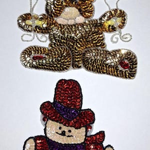 May include: Two sequined figures on a white background. The top figure is a teddy bear holding balloons. The bottom figure is a seated person wearing a hat. The figures are made of gold, red, green, blue, purple, and black sequins.