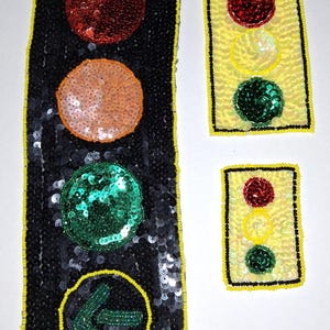 May include: Three sequined traffic light designs. One is black with red, orange, and green circles, and a yellow border. The other two are yellow with red, yellow, and green circles, and a black border. The largest has a green arrow.