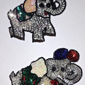May include: Two sequined elephant patches. The elephants are silver with black beaded outlines. One elephant holds sequined balloons. The elephants have colorful accents of pink, green, and red sequins.