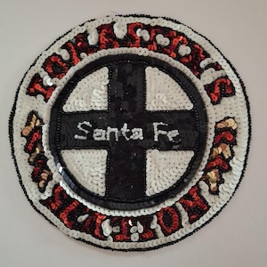 May include: Circular Santa Fe patch with a black cross design and the words "Santa Fe" in white. The patch is embellished with white, red, and gold sequins, and black beads. The patch is likely for a jacket or bag.