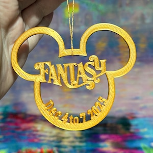 Puede incluir: Adorno dorado con forma de orejas de Mickey Mouse. La palabra "FANTASY" está en el centro, con "Dec 4 to 7 2025" debajo. El adorno está suspendido por un hilo dorado.