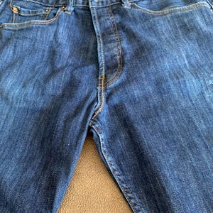 May include: Close-up of blue denim jeans with the "501" label visible. The jeans have classic five-pocket styling, a button fly, and copper-colored stitching. The fabric shows subtle fading and wear.