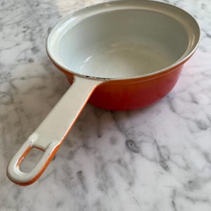 May include: A small, orange enameled saucepan with a white interior and handle. The handle is attached to the side of the pan. The pan is sitting on a white and gray marble surface. The pan is empty.