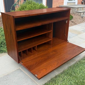 May include: A rectangular wooden wall-mounted cabinet with a rich brown finish. The unit features a drop-down door that serves as a flat surface, revealing an interior with multiple horizontal shelves and vertical dividers for organized storage.