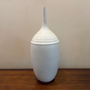 May include: A light gray ceramic vase with a textured surface and a long, slender neck. The vase has a rounded body and a lid with horizontal ridges. The vase is displayed on a dark wooden surface.