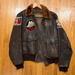 May include: A dark brown leather flight jacket featuring a brown faux fur collar, ribbed cuffs, and two front flap pockets. The jacket is adorned with various embroidered patches, including a hand graphic, national flags, and military insignia.