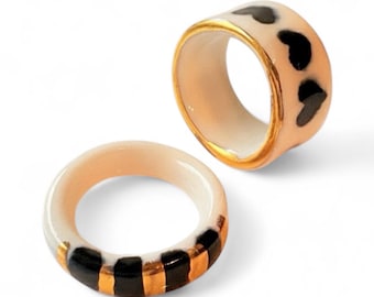 May include: Two ceramic rings with a smooth finish. One ring features a pattern of black hearts on a pale peach background with gold-toned edges. The second ring displays alternating vertical black and gold-toned stripes on a pale peach base.