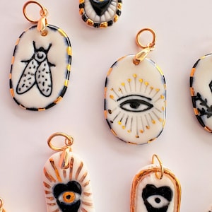 May include: A collection of oval-shaped ceramic pendants featuring hand-painted black and gold designs, including insects, eyes, and hearts. Each pendant has a gold-toned metal jump ring attached to the top for hanging on a necklace chain.