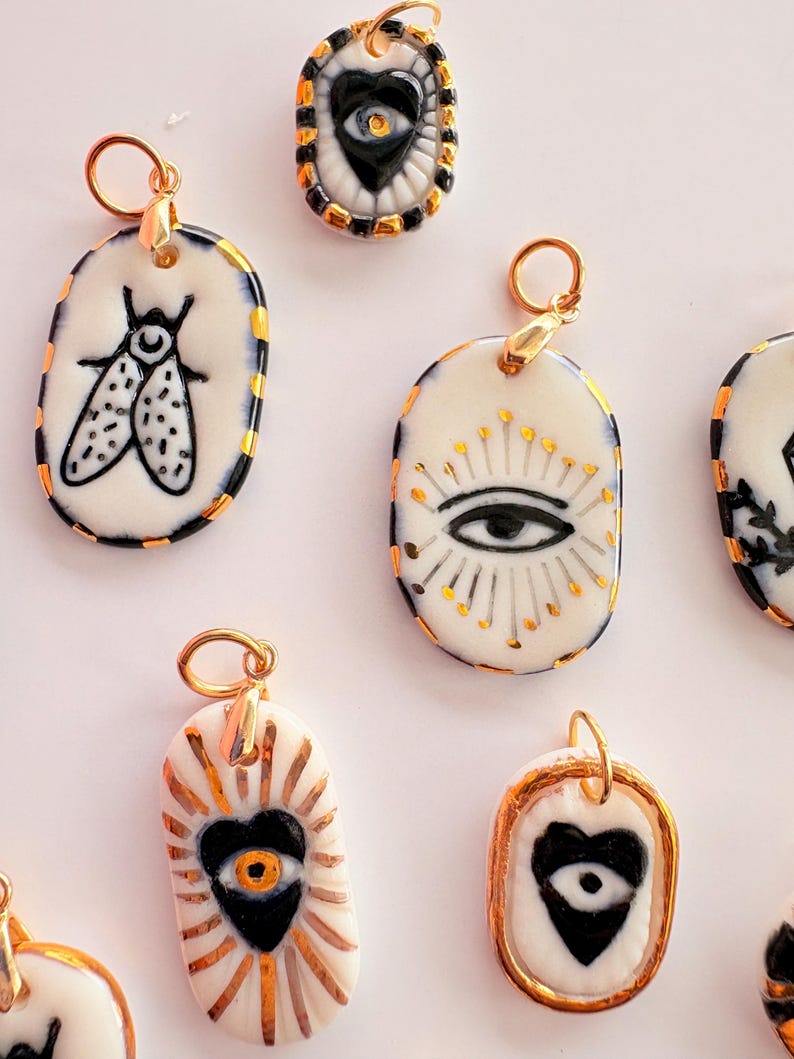 May include: A collection of oval-shaped ceramic pendants featuring hand-painted black and gold designs, including insects, eyes, and hearts. Each pendant has a gold-toned metal jump ring attached to the top for hanging on a necklace chain.