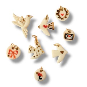 Peut inclure: Collection de breloques en céramique crème avec des détails dorés. Les breloques présentent divers motifs, notamment des colombes, des cœurs, des nœuds et des motifs floraux. Chaque breloque a une boucle dorée pour la fixation.