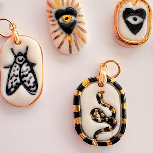 May include: A collection of oval-shaped ceramic pendants featuring hand-painted black and white designs including a moth, a heart with an eye, and a coiled snake. Each pendant is accented with metallic gold details and includes a gold jump ring.