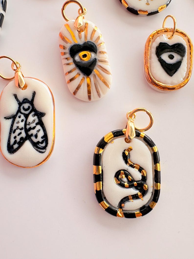 May include: A collection of oval-shaped ceramic pendants featuring hand-painted black and white designs including a moth, a heart with an eye, and a coiled snake. Each pendant is accented with metallic gold details and includes a gold jump ring.