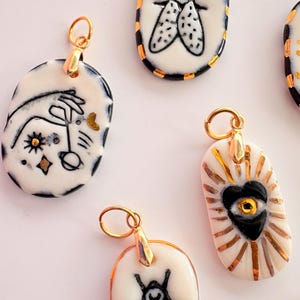 May include: A collection of oval-shaped ceramic pendants featuring hand-painted black ink illustrations of insects, celestial motifs, and a central heart with an eye, all accented with metallic gold detailing and attached to gold-toned metal jump rings.