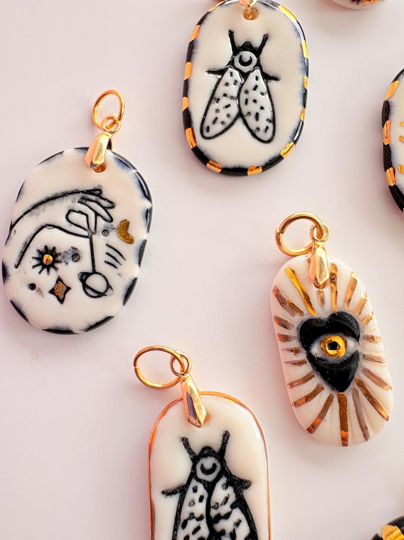 May include: A collection of oval-shaped ceramic pendants featuring hand-painted black ink illustrations of insects, celestial motifs, and a central heart with an eye, all accented with metallic gold detailing and attached to gold-toned metal jump rings.