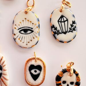 May include: A collection of oval and circular ceramic pendants featuring hand-painted black and gold designs, including an eye with rays, a crystal, a heart with an eye, and a skull. Each pendant has a gold-toned metal jump ring attached.