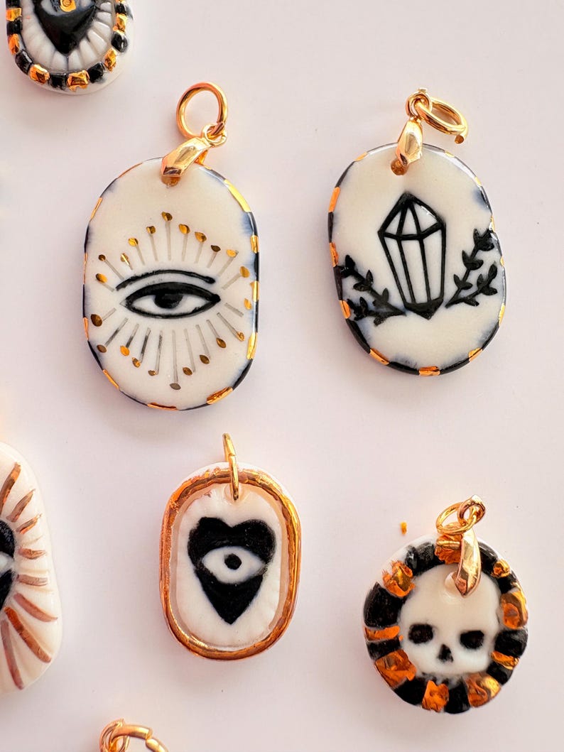 May include: A collection of oval and circular ceramic pendants featuring hand-painted black and gold designs, including an eye with rays, a crystal, a heart with an eye, and a skull. Each pendant has a gold-toned metal jump ring attached.