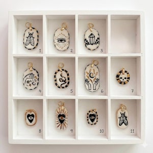 May include: Eleven oval and circular ceramic pendants with gold-toned jump rings, each featuring black ink illustrations including a moth, eye, crystal, hand, snake, moon, skull, and heart. The pendants are arranged in a white grid display case.