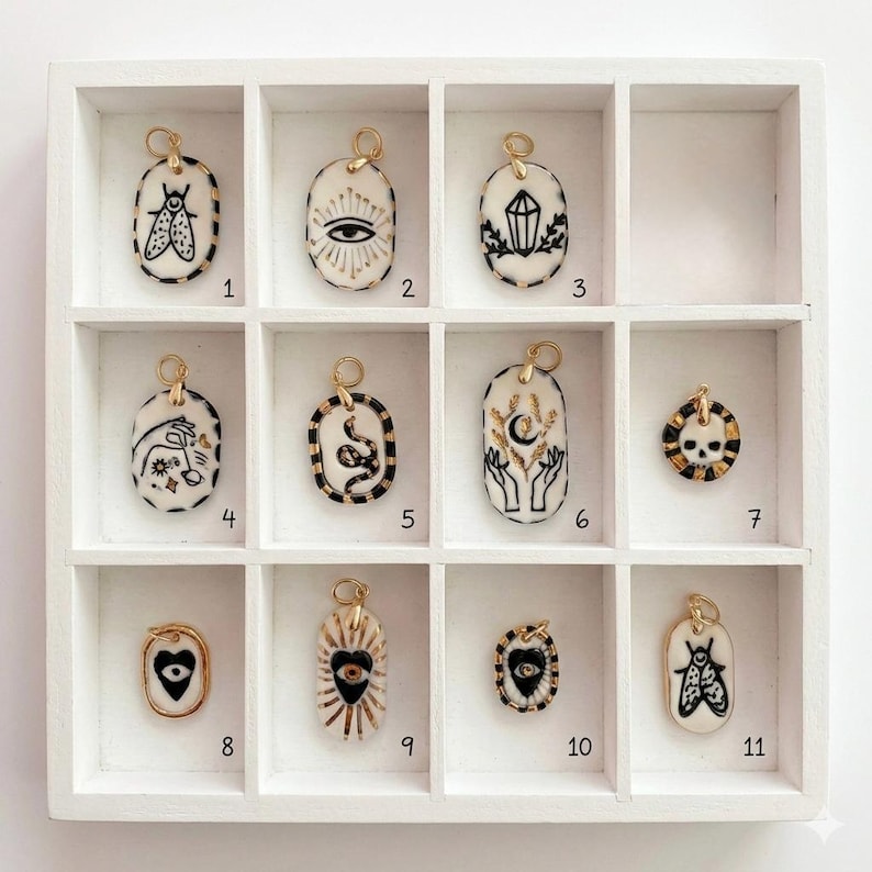 May include: Eleven oval and circular ceramic pendants with gold-toned jump rings, each featuring black ink illustrations including a moth, eye, crystal, hand, snake, moon, skull, and heart. The pendants are arranged in a white grid display case.