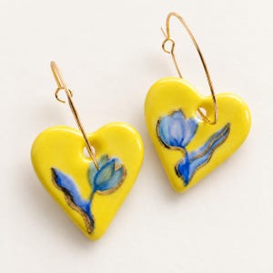 Könnte beinhalten: Ein Paar herzförmige Ohrringe mit einer leuchtend gelben Glasur und einem handgemalten blauen Tulpenmotiv. Jeder herzförmige Anhänger hängt an einem dünnen, goldfarbenen Metallreifen und bildet so ein zusammenhängendes Set an Schmuck.