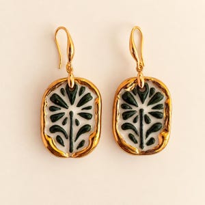 Puede incluir: Un par de pendientes colgantes de tono dorado con un colgante ovalado que presenta un centro de esmalte blanco y un motivo floral verde oscuro. Los pendientes tienen un borde de metal dorado pulido y un cierre de gancho curvado.