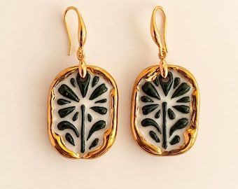 May include: A pair of gold-toned drop earrings featuring an oval-shaped pendant with a white enamel centre and a dark green floral motif. The earrings have a polished gold metal border and a curved hook closure for secure attachment to the ear.
