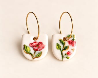 May include: A pair of hoop earrings featuring white, semi-circular ceramic pendants. Each pendant is hand-painted with a red floral design, green leaves, and gold accents. The thin gold-toned metal hoops attach through a hole at the top.