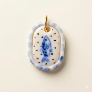 May include: Oval pendant featuring a blue fish design. The pendant has a white base with blue and gold detailing. A gold-coloured loop is at the top for hanging. The pendant is likely ceramic or porcelain.