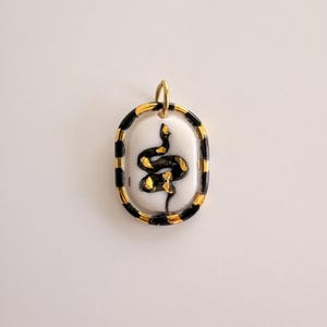 Puede incluir: Colgante ovalado con una fianza dorada, con un diseño de serpiente negra y dorada sobre un fondo blanco. El borde del colgante es a rayas negras y doradas. Una joya única.