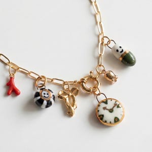 Puede incluir: Collar con colgantes dorados con una variedad de dijes. Los dijes incluyen un coral rojo, una esfera a rayas blancas y negras con una cara sonriente, un lazo dorado, un reloj, una píldora y una pequeña campana. Fondo blanco.