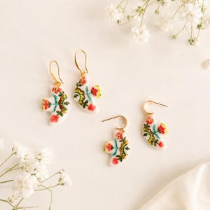 Puede incluir: Dos pares de pendientes colgantes con forma de pájaro, con dijes de cerámica blanca y motivos florales pintados a mano en rojo, verde y dorado. Cada pendiente cuelga de un fino gancho de metal de tono dorado con silueta aviar.