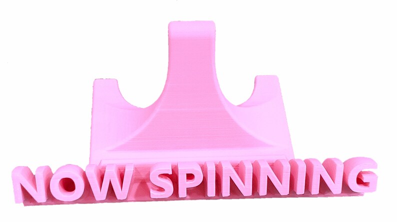 K&ouml;nnte beinhalten: Ein rosa 3D-gedruckter Telefonst&auml;nder mit dem Schriftzug "NOW SPINNING" auf der Vorderseite.