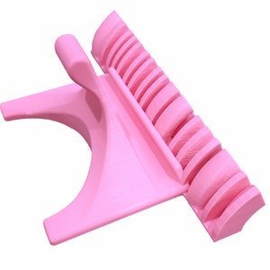 K&ouml;nnte beinhalten: Ein rosa Telefonst&auml;nder aus Kunststoff mit integriertem Ladedock. Der St&auml;nder hat eine gebogene Basis und einen erh&ouml;hten Bereich mit Schlitzen zum Halten mehrerer Telefone.
