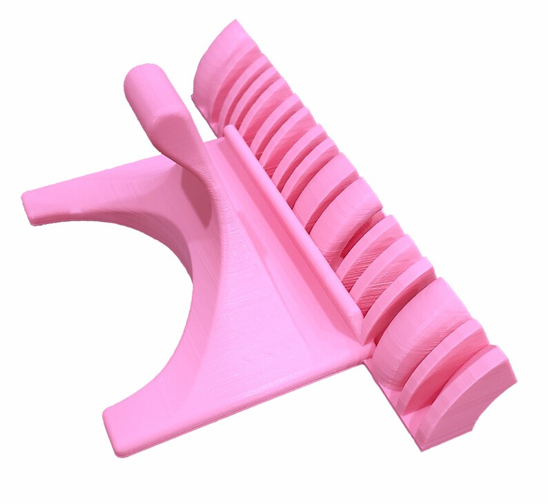 K&ouml;nnte beinhalten: Ein rosa Telefonst&auml;nder aus Kunststoff mit integriertem Ladedock. Der St&auml;nder hat eine gebogene Basis und einen erh&ouml;hten Bereich mit Schlitzen zum Halten mehrerer Telefone.