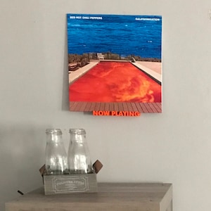 May include: A Red Hot Chili Peppers album cover titled "Californication" featuring a swimming pool with red water and a blue sky. The album cover is on a wall with the words "NOW PLAYING" in orange letters below it.