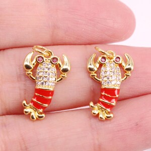 Puede incluir: Dos colgantes dorados con forma de langosta, con colas de esmalte rojo y detalles de cristal transparente en el cuerpo. Los colgantes de langosta tienen ojos de gema roja y están diseñados con un lazo para colgar.