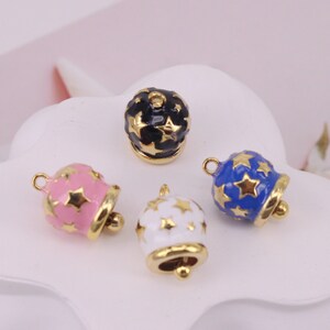 Puede incluir: Cuatro pequeñas campanas doradas con esmalte de diferentes colores y estrellas doradas. Las campanas son rosa, azul, blanca y negra.