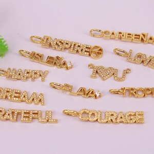 Könnte beinhalten: Eine Sammlung von goldfarbenen Charms mit verschiedenen Wörtern und Symbolen, darunter "LOVE", "DREAM", "HAPPY", "INSPIRE", "COURAGE", "GRATEFUL", "PLAYA", "TROPICAL", "ISLEÑA", "CARIBENA" und "I LOVE YOU". Jeder Charm ist mit kleinen, klaren Strasssteinen verziert.
