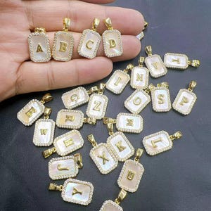 May include: A collection of 26 gold-tone pendants with white mother-of-pearl inlays and clear rhinestones. Each pendant features a different letter of the alphabet.