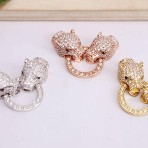 Könnte beinhalten: Drei dekorative Schmuckverschlüsse in Silber-, Roségold- und Goldtönen. Jeder Verschluss zeigt zwei Tierköpfe, die mit kleinen, klaren Kristallen besetzt sind und durch einen Ring verbunden sind. Die Verschlüsse werden auf einer weißen Oberfläche präsentiert.