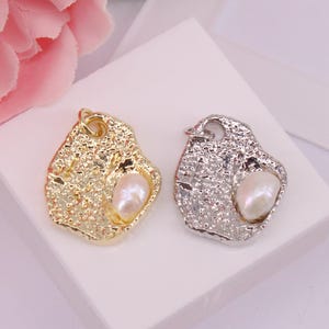 以下が含まれることがあります： 金色のものと銀色のもの、2つのペンダントチャーム。それぞれがテクスチャ加工された不規則な形状をしています。白い涙型の真珠が各チャームに収められています。チャームは白い表面に表示されています。