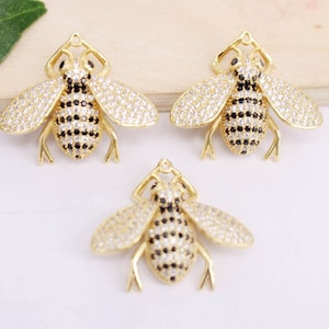 May include: Three gold-colored bee charms. The bee charms have clear and black crystal accents on the wings and body. The charms are on a white surface.