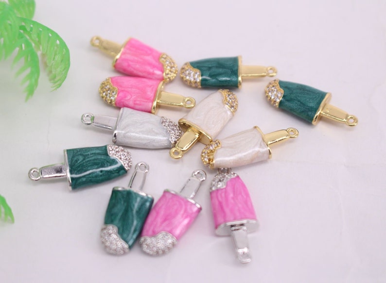 Puede incluir: Surtido de abalorios de paleta de colores con detalles dorados y plateados. Los abalorios est&aacute;n hechos de esmalte y presentan una variedad de colores, incluyendo rosa, verde y blanco. Cada abalorio tiene un bucle en la parte superior para sujetarlo a un collar, pulsera u otras joyas.