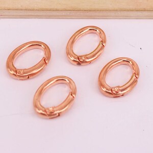 May include: Four oval-shaped, rose gold-colored clasps. Each clasp has a hinged design for easy opening and closing. The clasps are arranged on a light pink surface, with a wooden element in the background.