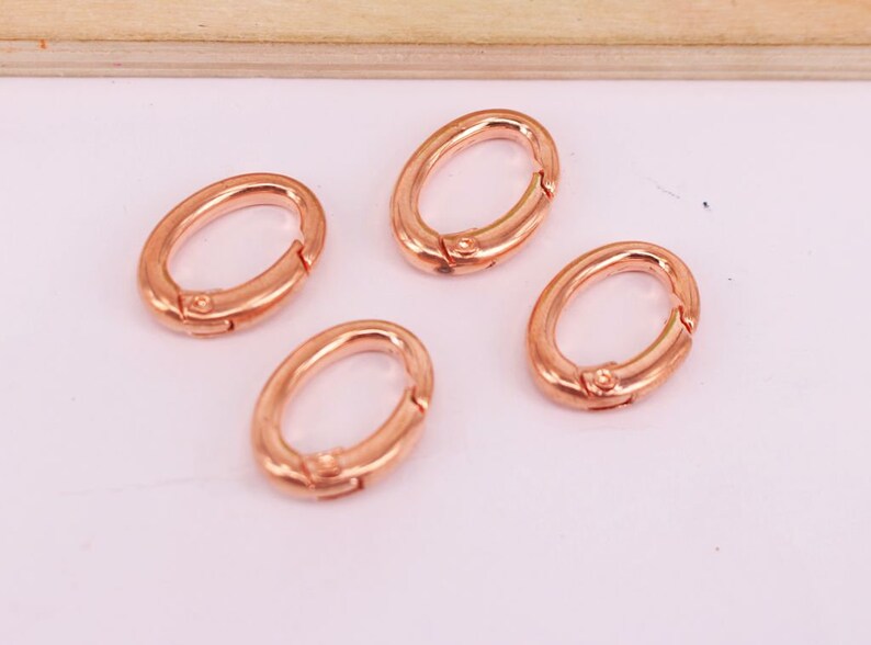 May include: Four oval-shaped, rose gold-colored clasps. Each clasp has a hinged design for easy opening and closing. The clasps are arranged on a light pink surface, with a wooden element in the background.