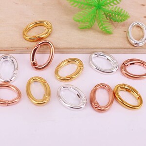May include: An assortment of oval-shaped metal clasps in gold, silver, and rose gold. The clasps are open, showcasing their hinged design. The background is a light pink surface with a wooden element and a green plastic plant.